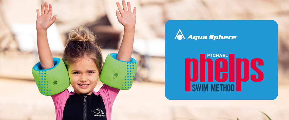 Kleines Mädchen im Neopren-Anzug und mit grünen Schwimmflügeln hebt die Hände über den Kopf.